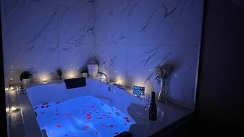 A modern bathroom with a marble wall, a hot tub with rose petals, a digital control panel, and a bottle of champagne.
