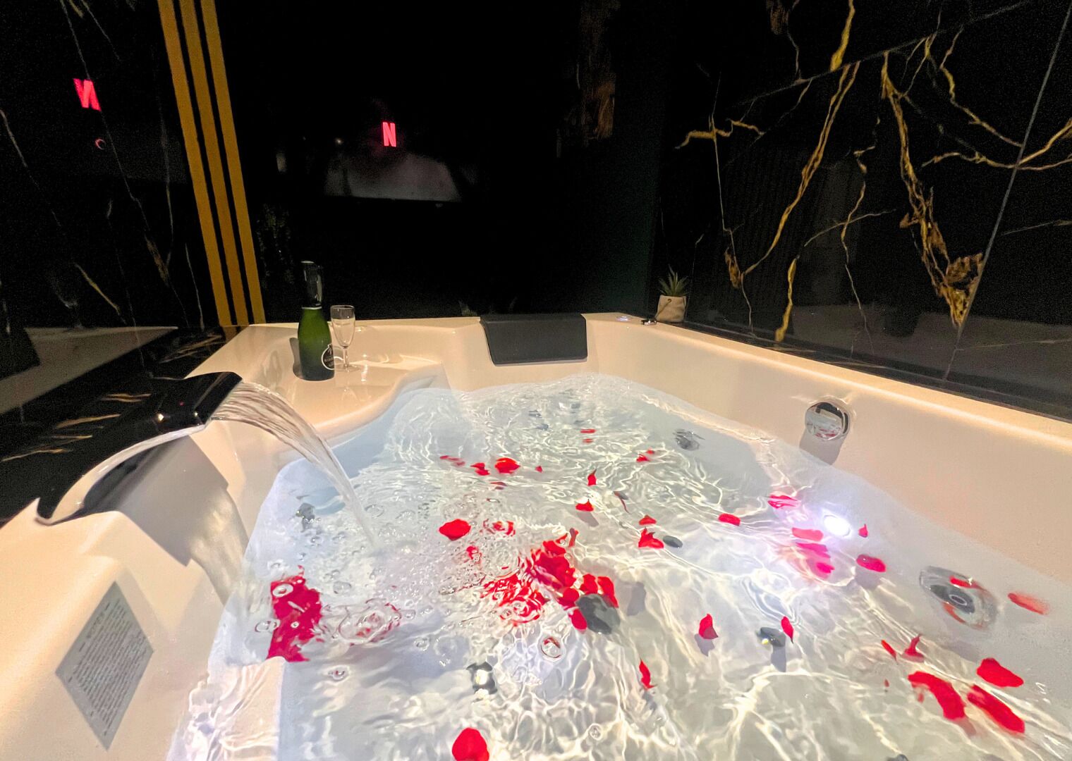 Una bañera de hidromasaje con pétalos de rosa flotando en el agua, una copa de vino y una botella a un lado.