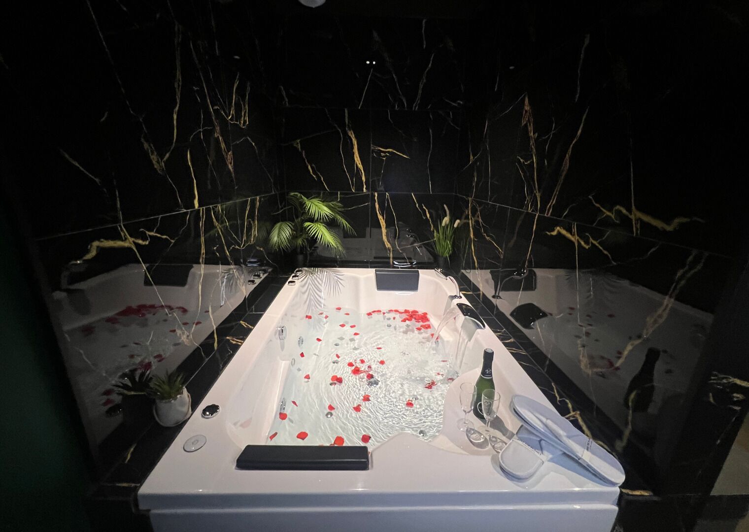 A bathtub with rose petals, a bottle, and glasses, surrounded by plants and a dark background.