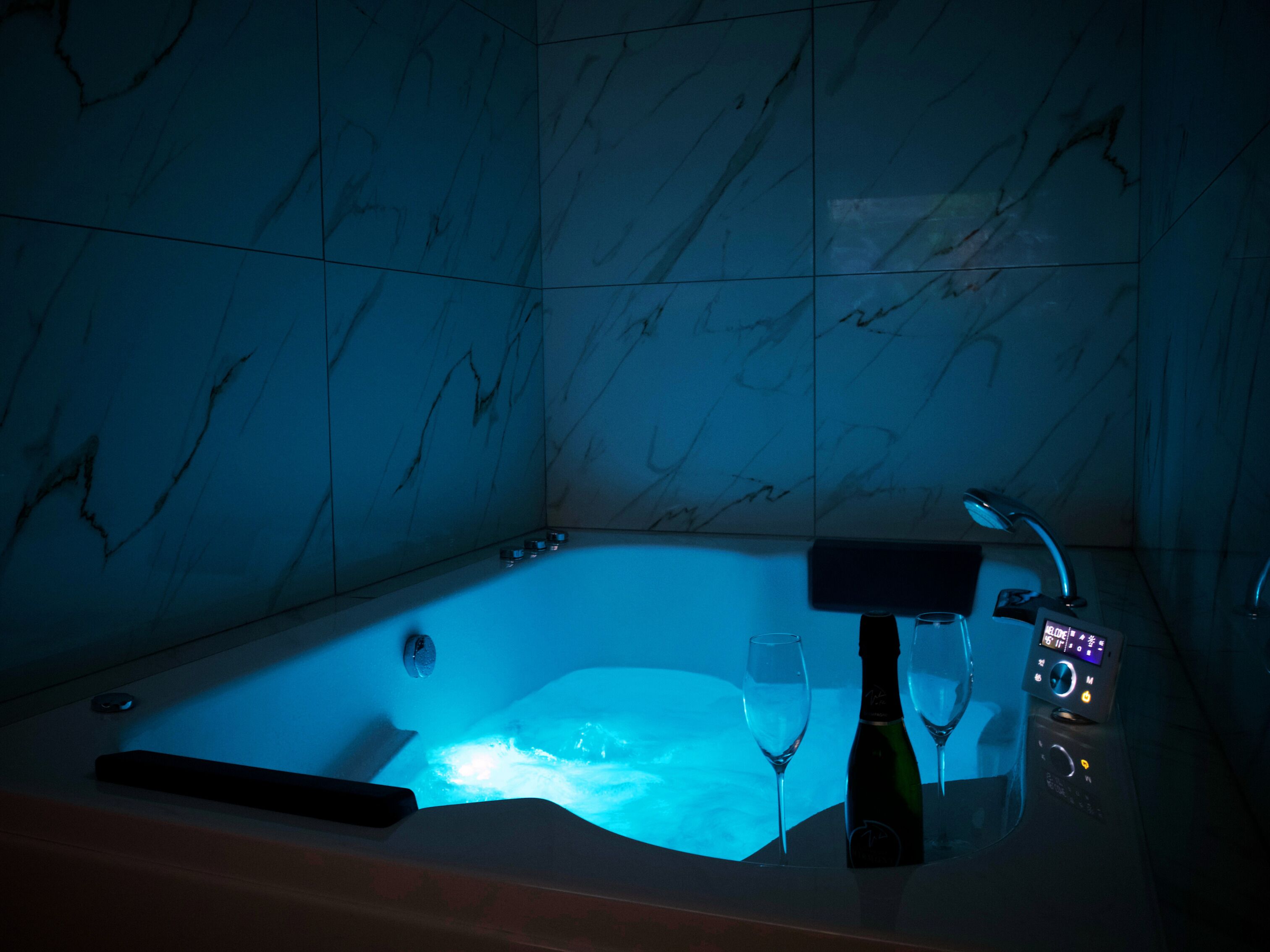 Une pièce faiblement éclairée avec un mur de marbre, une baignoire, une bouteille de champagne et deux verres.