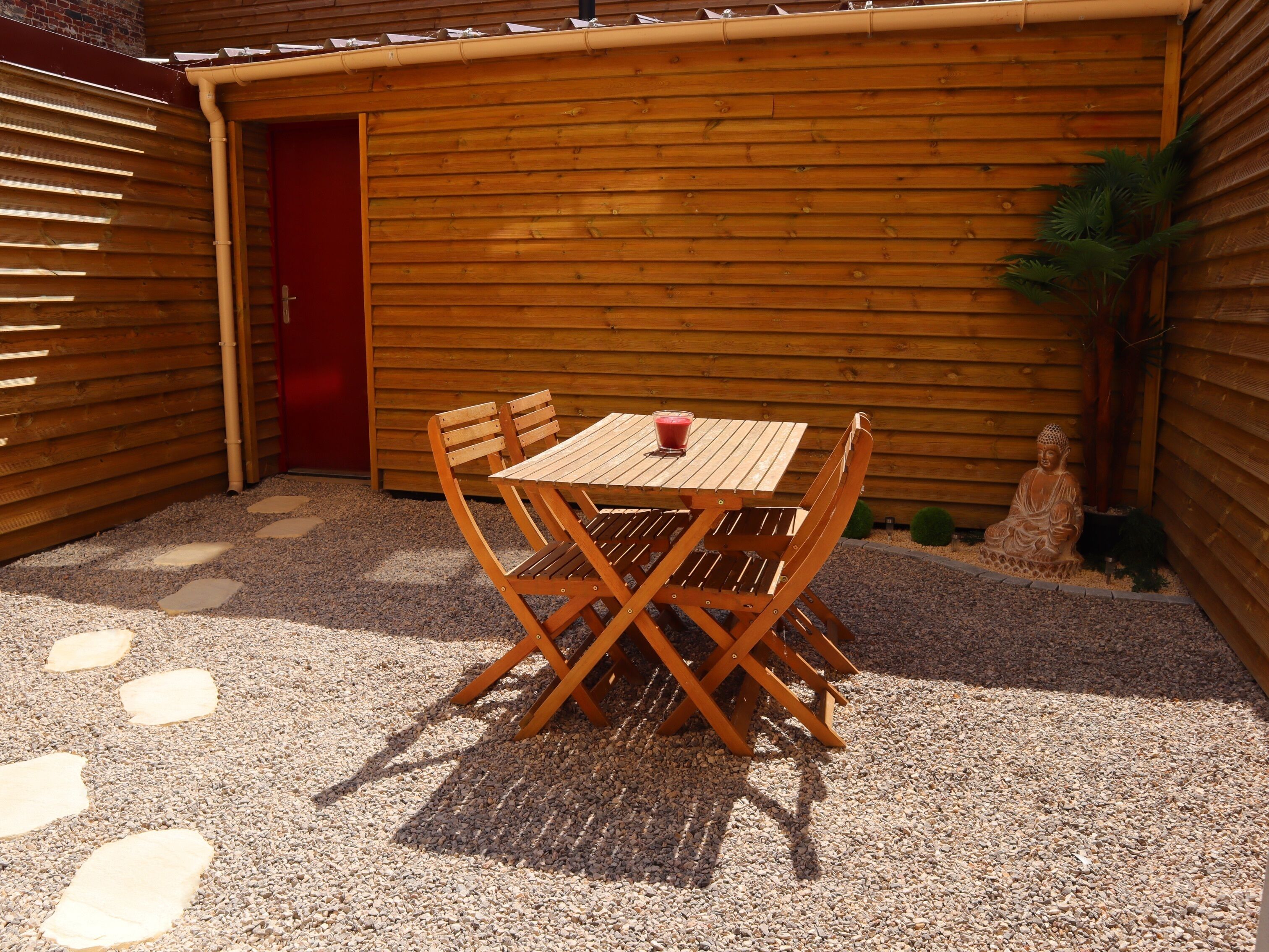 A wooden table and chairs set on a gravel patio with a red door and wooden walls.