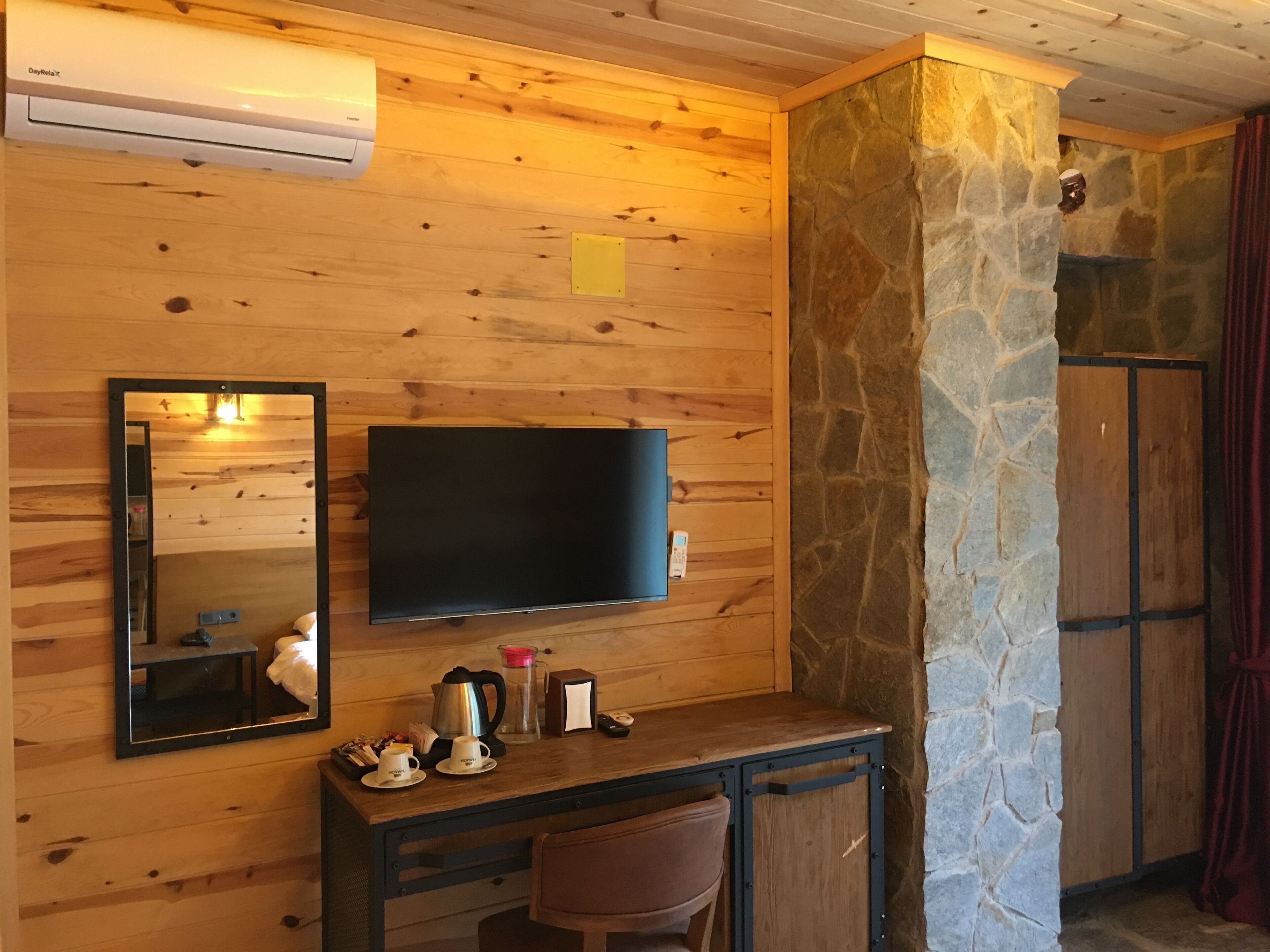 A wooden-paneled room with a mounted TV, a stone wall, a wooden desk with a chair, and a stone fireplace.