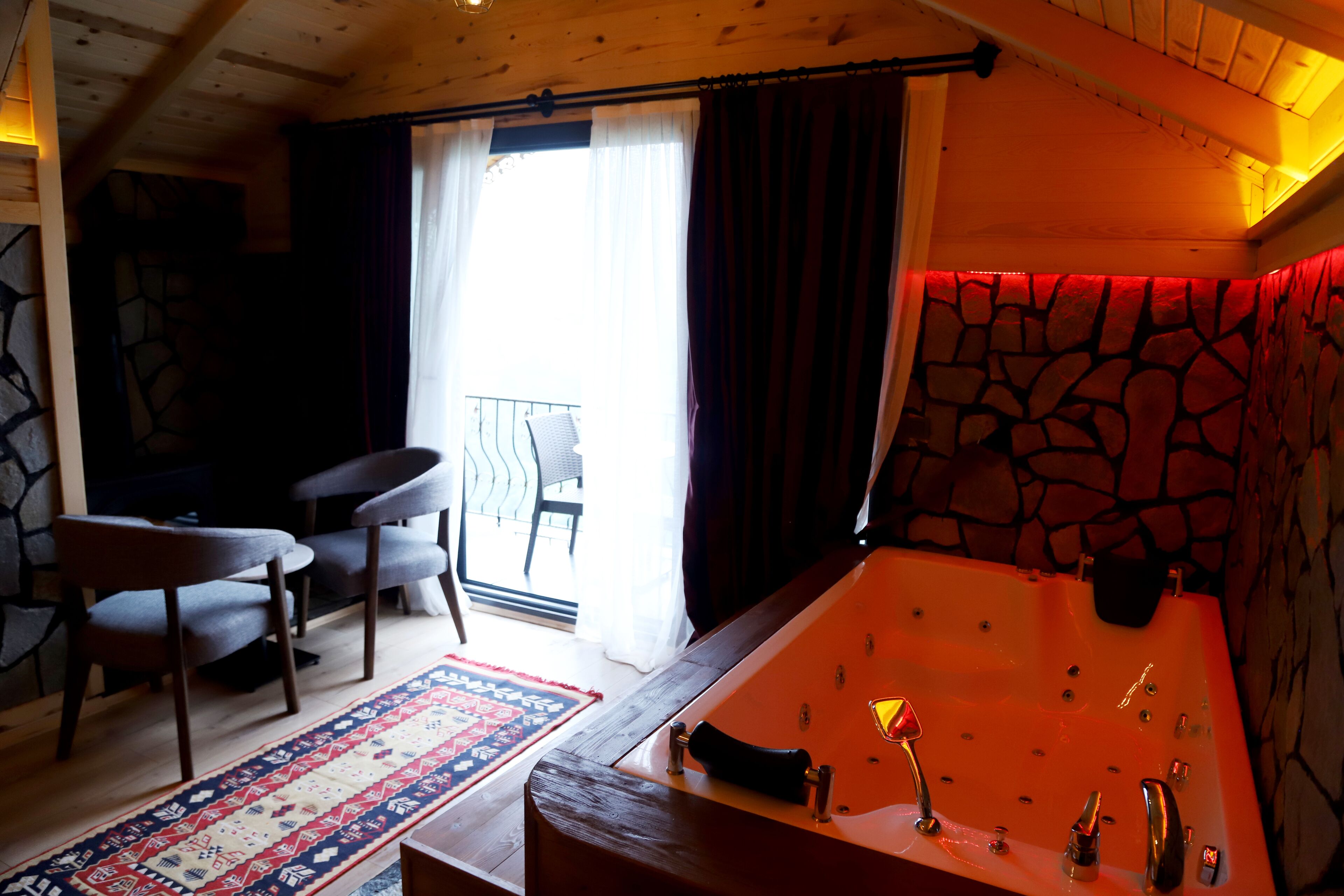 A room with a stone wall, a large bathtub, a wooden ceiling, and a view of the outside through a window.