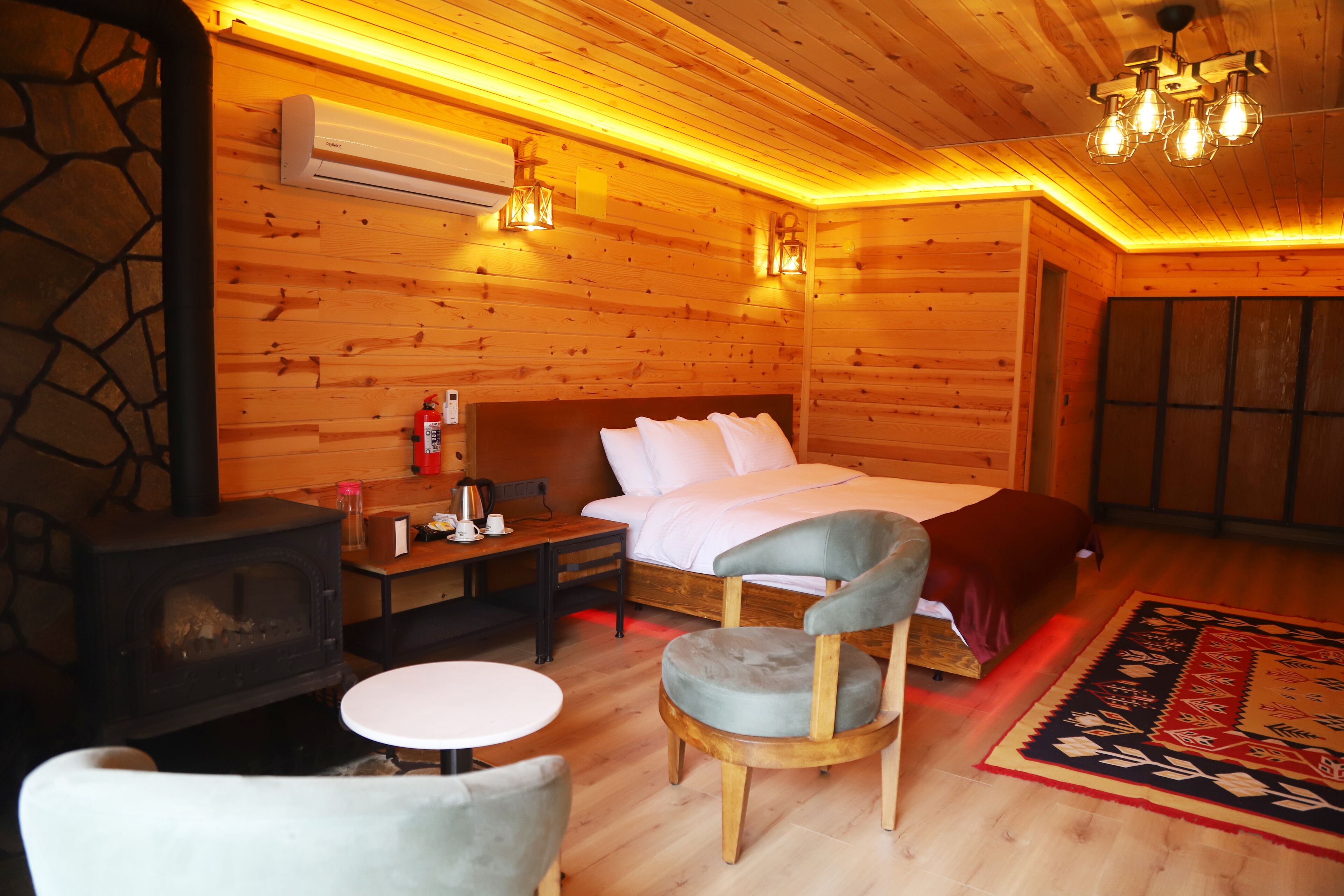 A cozy wooden cabin room with a bed, a small table, a chair, a fireplace, and a rug.