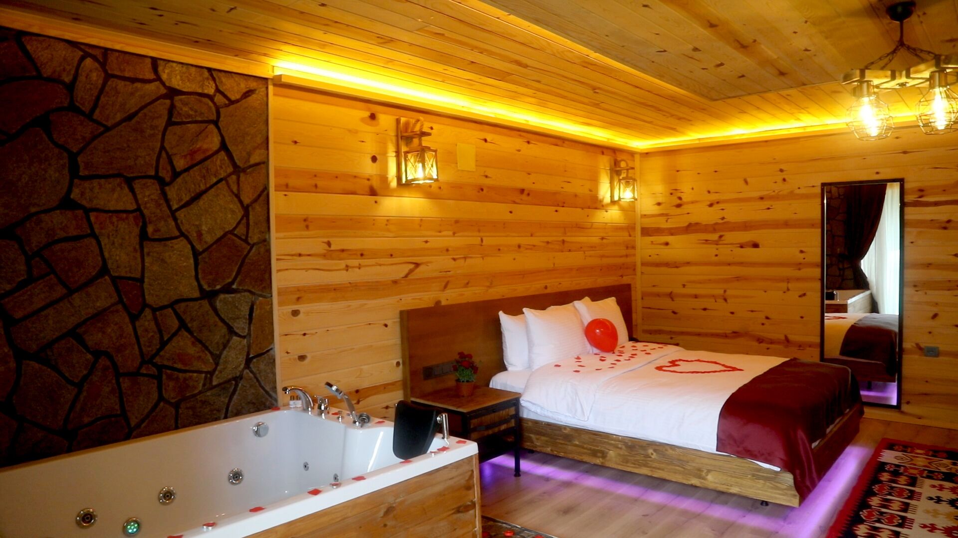 A wooden cabin room with a stone wall, a large bed with a red heart decoration, a wooden nightstand, a red chair, a large bathtub, and a mirror on the wall.