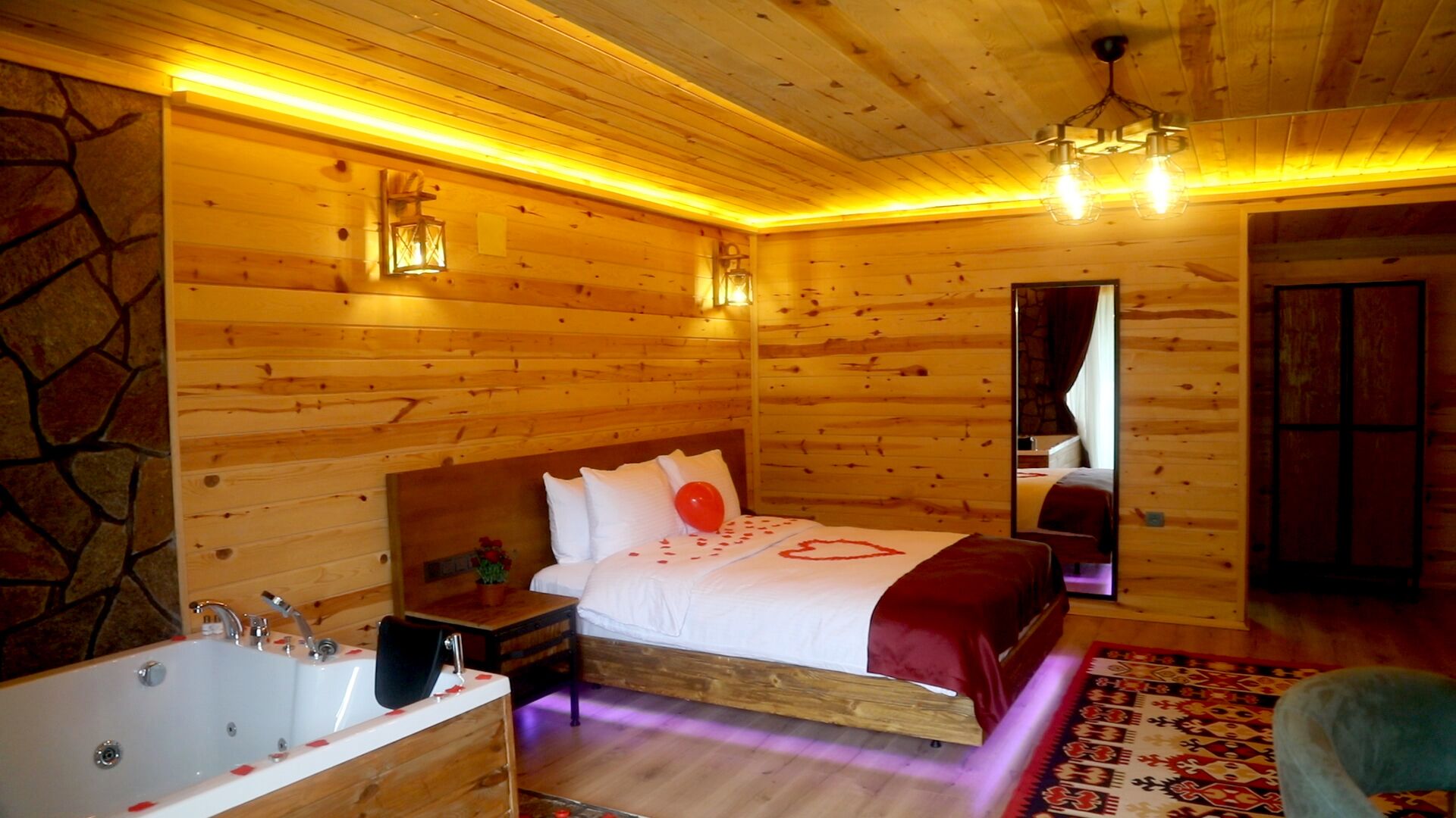 Un dormitorio de cabaña de madera con una cama, una mesita de noche, una colcha roja, un baño con bañera y una pared de piedra.