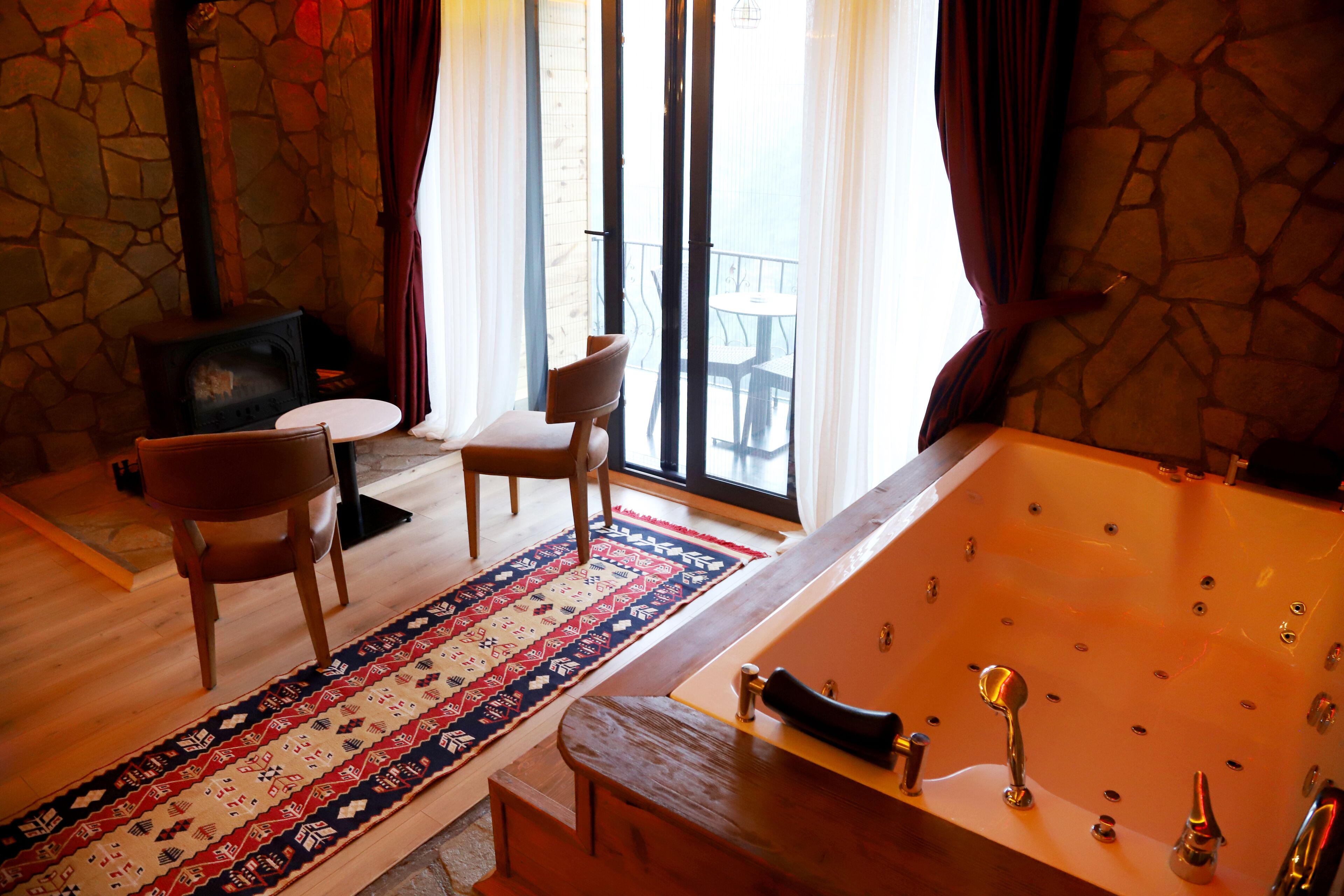 A room with a stone wall, a large bathtub, a wooden chair, a small table, and a patterned rug.