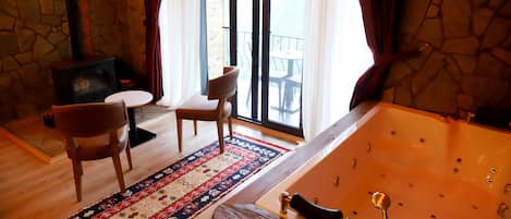 A room with a stone wall, a large bathtub, a wooden chair, a small table, and a patterned rug.