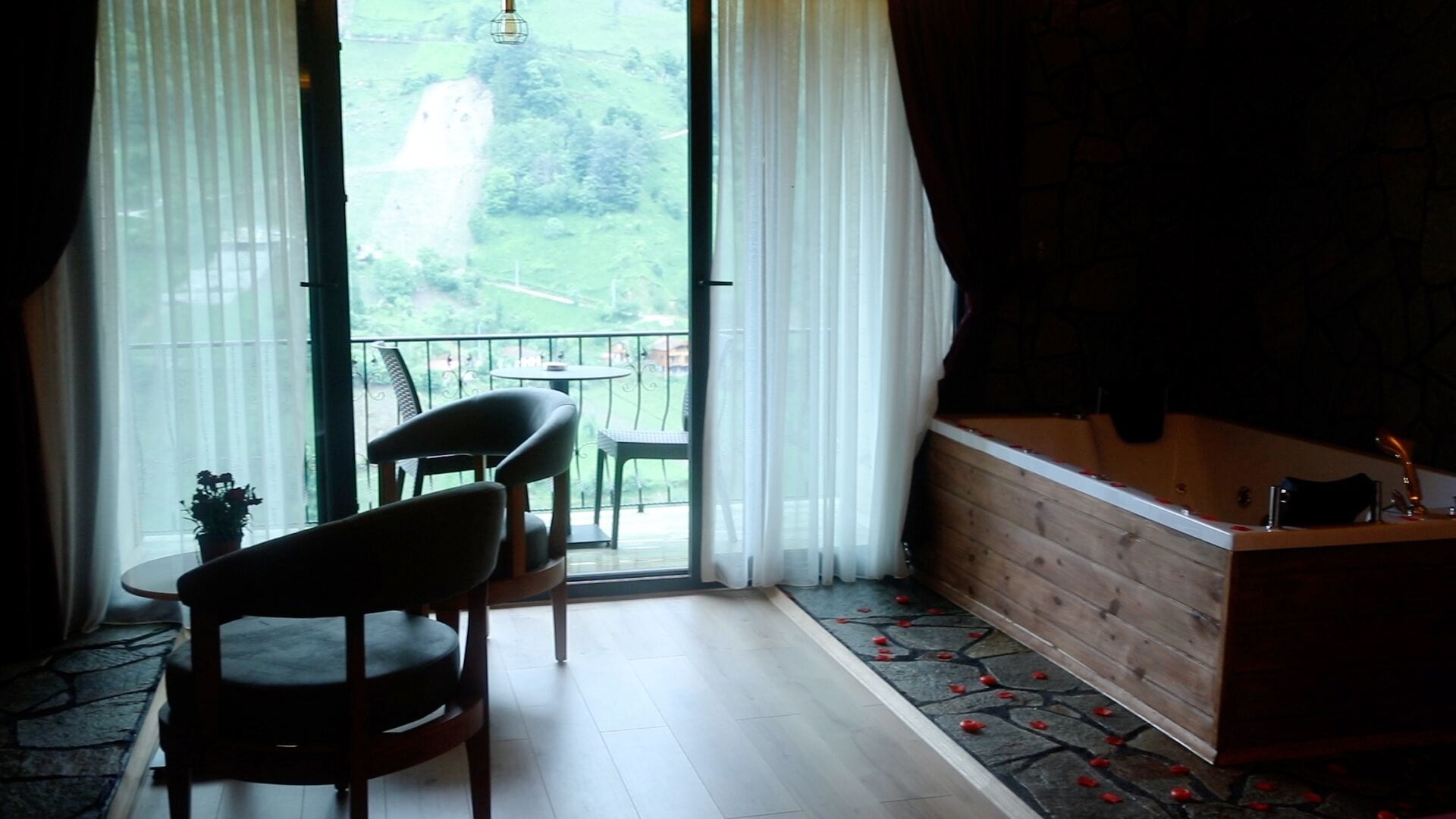 Quarto com piso de madeira, uma pequena mesa com cadeiras, um banco de madeira, uma banheira e vista para uma paisagem verde através de uma porta de vidro.