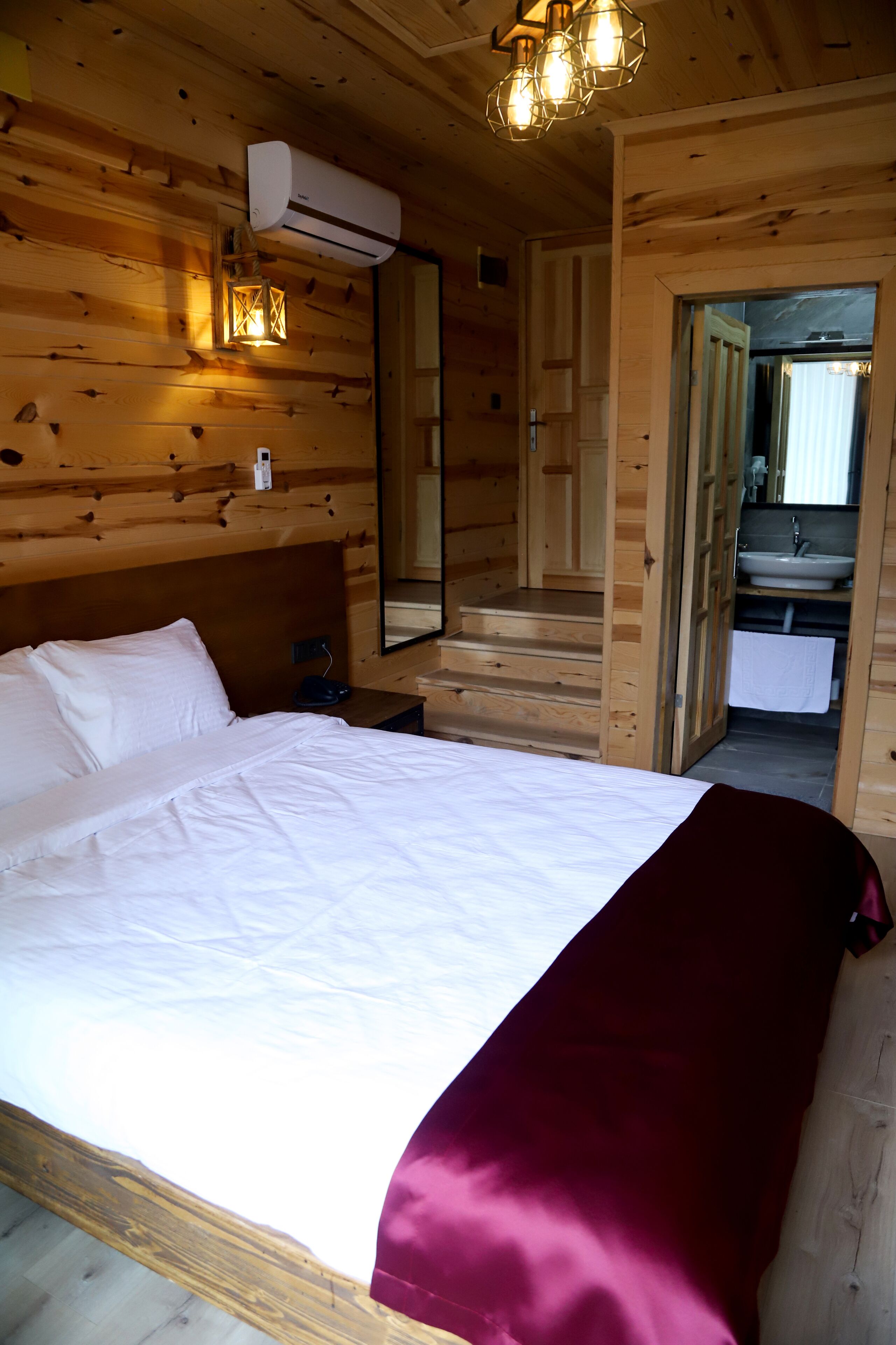 A wooden cabin bedroom with a bed, a nightstand, a mirror, and a bathroom visible through an open door.