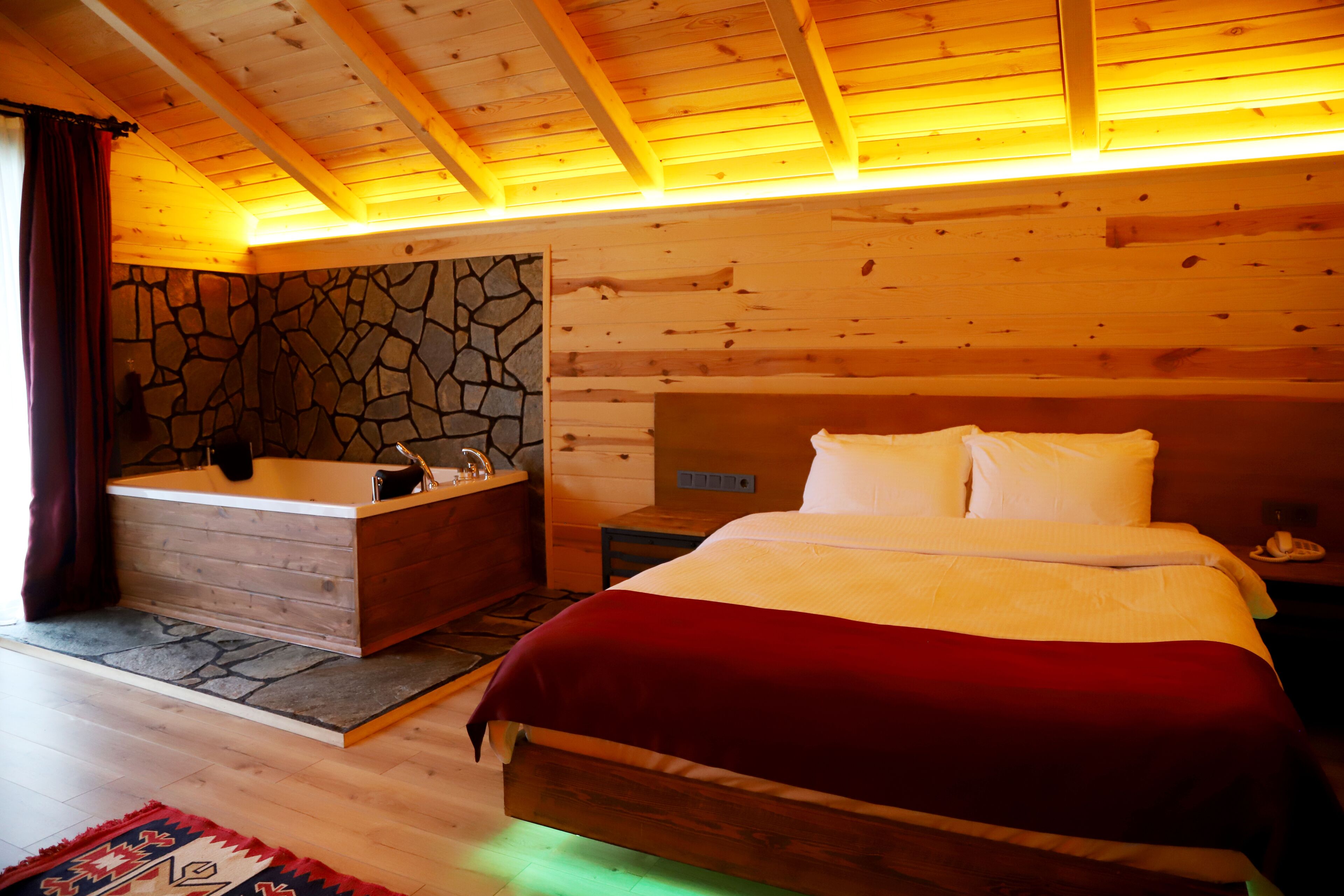 A wooden cabin bedroom with a stone wall, a wooden bed, a nightstand, and a bathroom with a bathtub.