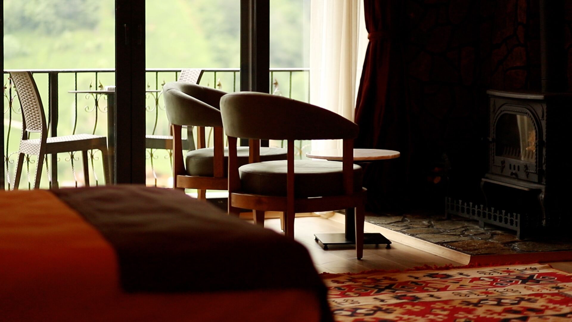 A room with a bed, two chairs, a small table, a fireplace, and a view of greenery outside.