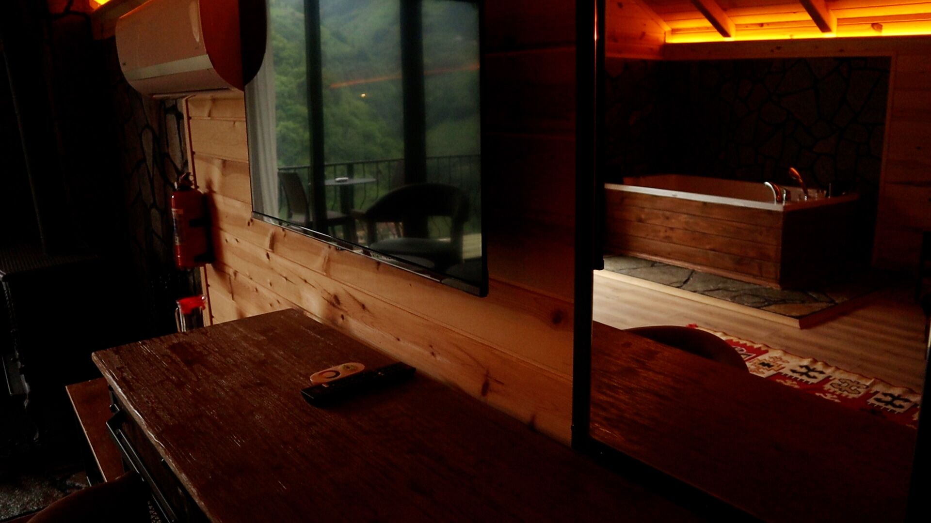 Interior de una cabaña de madera con un ventanal grande, una mesa de madera y un pequeño rincón de descanso.