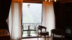 A cozy room with a wooden floor, a fireplace, a table with chairs, and a view of a mountainous landscape through a large window with curtains. - Yamaçda Suit Otel (Camlihemsin)