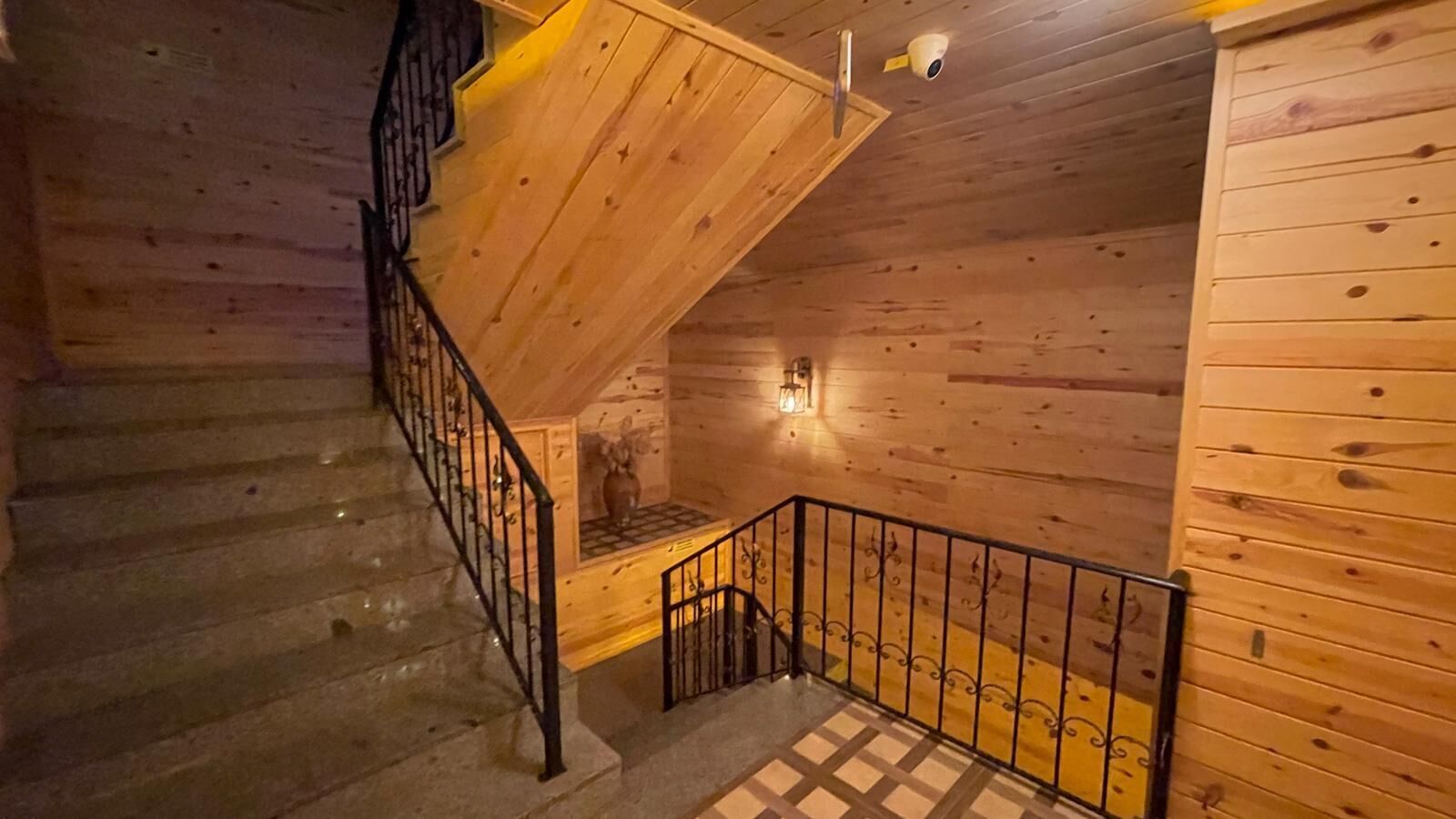 A wooden staircase with a metal railing, a wall-mounted light fixture, and a wooden floor with a geometric pattern.