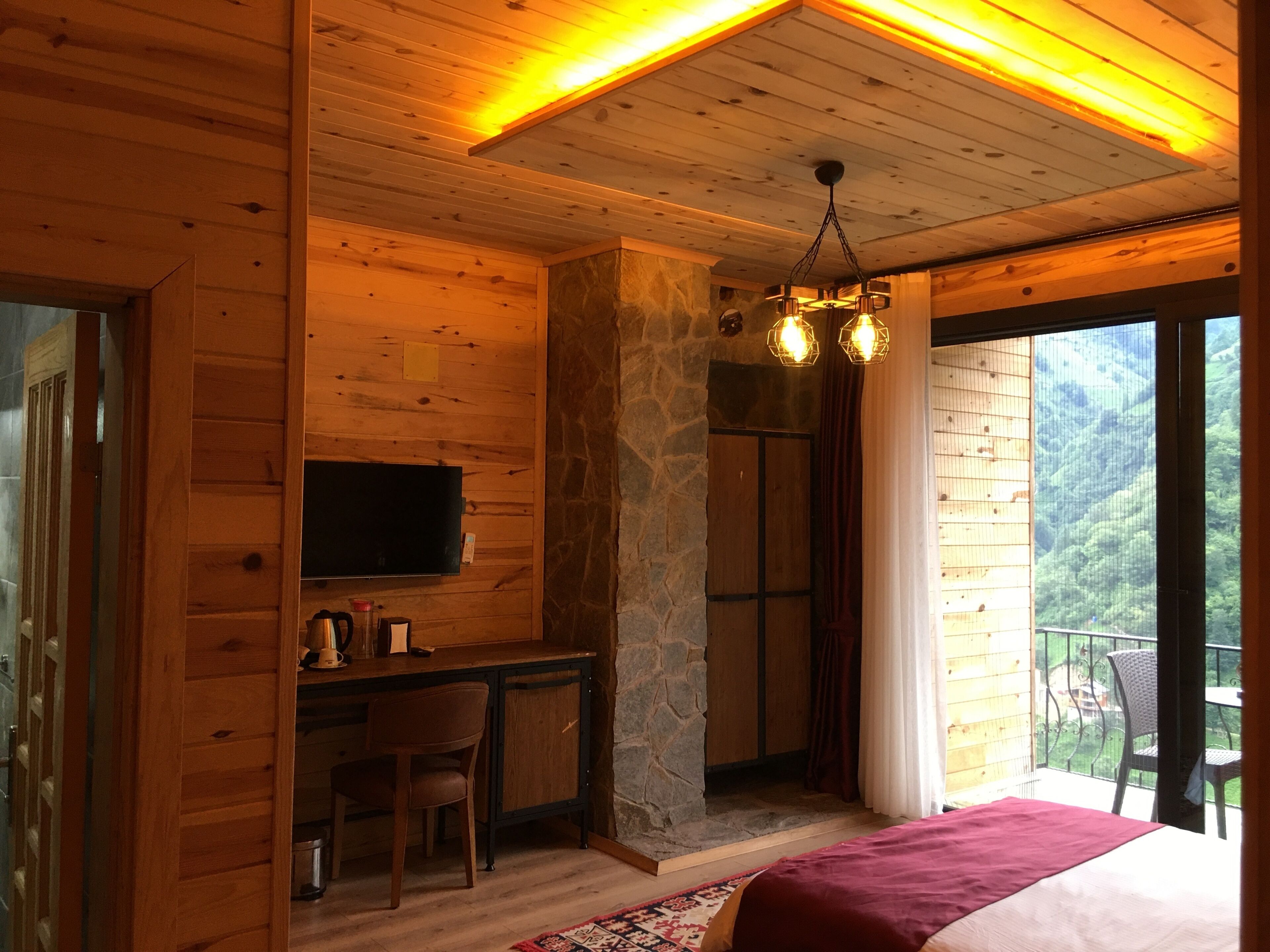 A wooden cabin bedroom with a stone fireplace, a desk with a chair, a bed, and a balcony with a view of the forest.