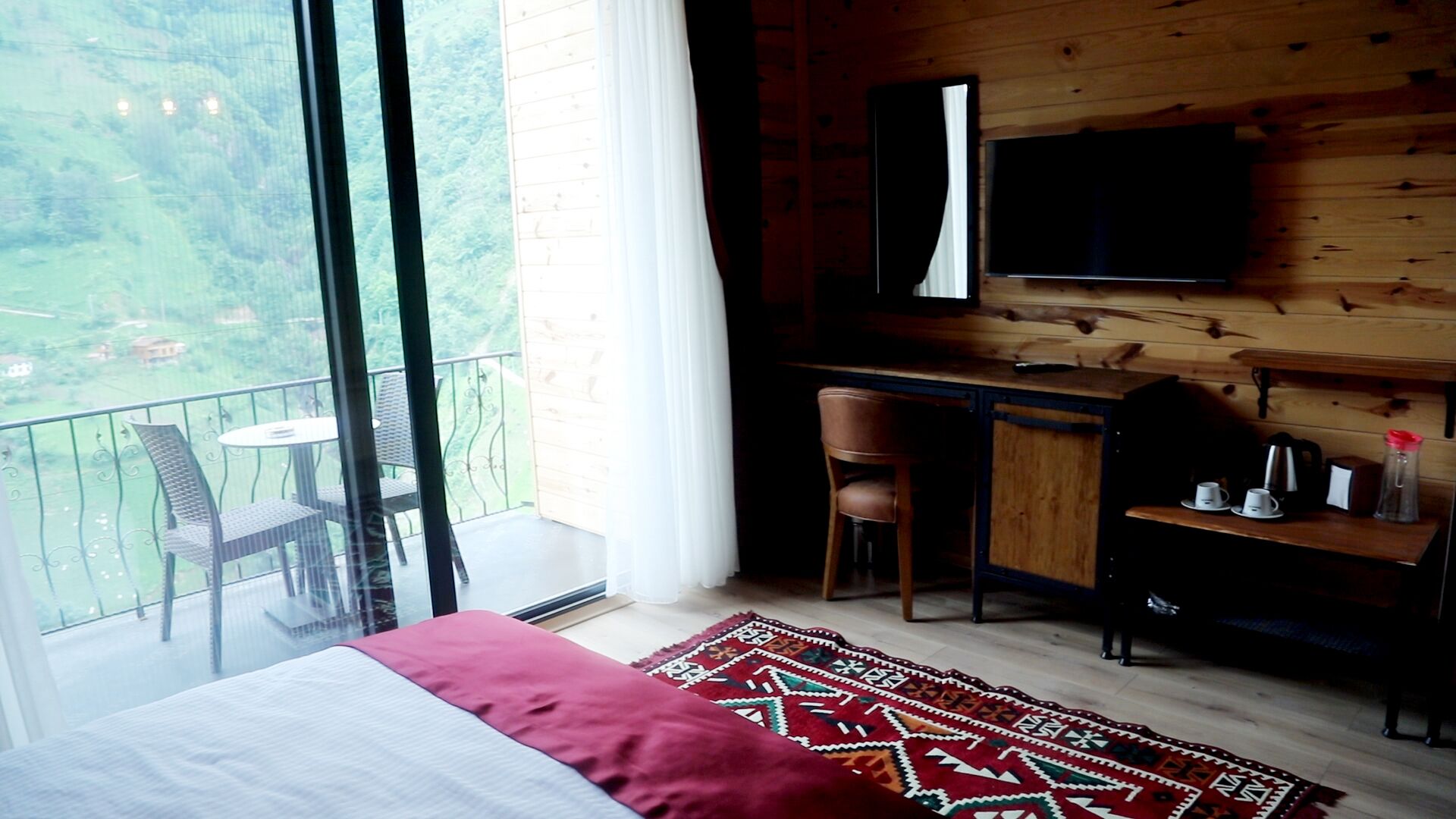 Un dormitorio con una cama grande, un escritorio con silla, un televisor montado en la pared y un balcón con mesa y sillas.
