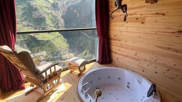 A wooden cabin interior with a large window, a wooden chair, a round bathtub, and a hanging light fixture. - Yamaçda Suit Otel (Camlihemsin)