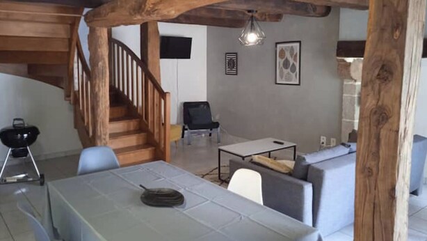 Ein rustikaler Innenraum mit freiliegenden Holzbalken, einer Treppe, einem Essbereich mit Tisch und Stühlen sowie einem Wohnbereich mit Sofa und Fernseher.