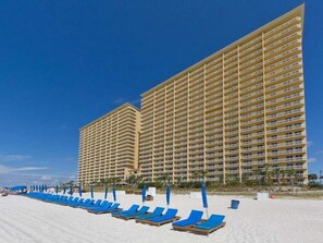 Un hotel con varios pisos y balcones, una playa con tumbonas y un cielo azul despejado.