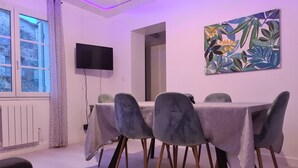 Un coin repas moderne avec une table dressée pour quatre personnes, entourée de chaises grises aux pieds en bois. Un téléviseur à écran plat est fixé au mur, et un tableau abstrait aux couleurs vives est visible.