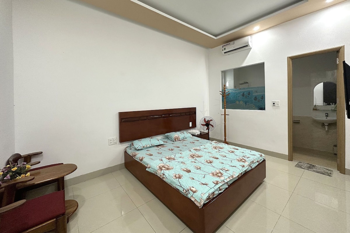 Quarto com uma cama, mesa de cabeceira de madeira, escrivaninha com cadeira, ventilador e janela com vista para o exterior.