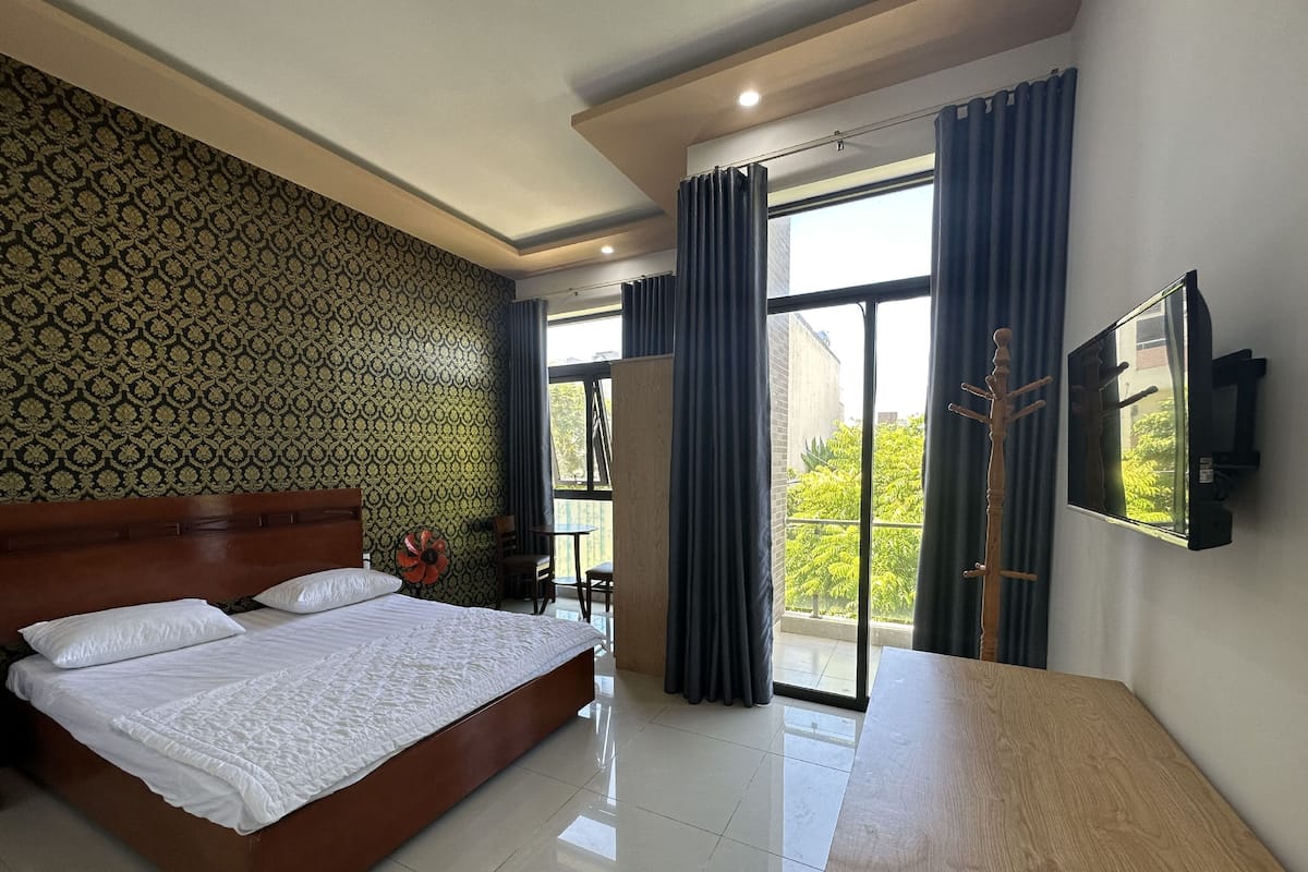 Quarto de hotel com uma cama grande, uma mesa de cabeceira de madeira, uma TV de tela plana e uma varanda com vista para árvores.