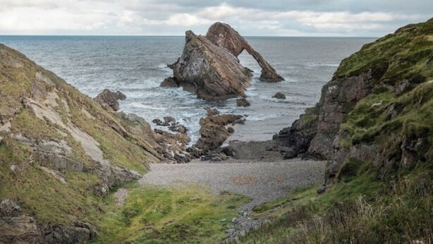 A coastal landscape with a prominent rock formation, a narrow path leading to the sea, and grassy hills. - Pretty cottage on Moray coast: walks & whisky (Buckie)