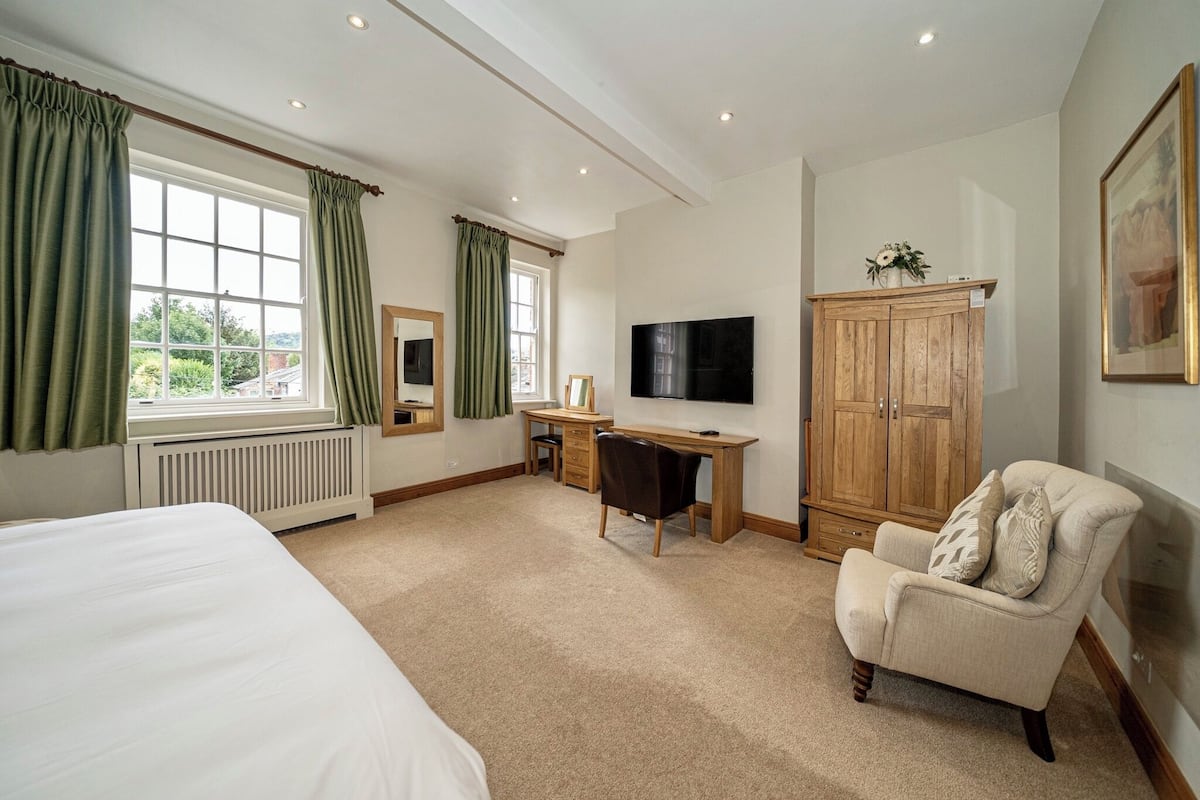 A hotel room with a large bed, a desk with a chair, a television, a wardrobe, and a window with curtains.