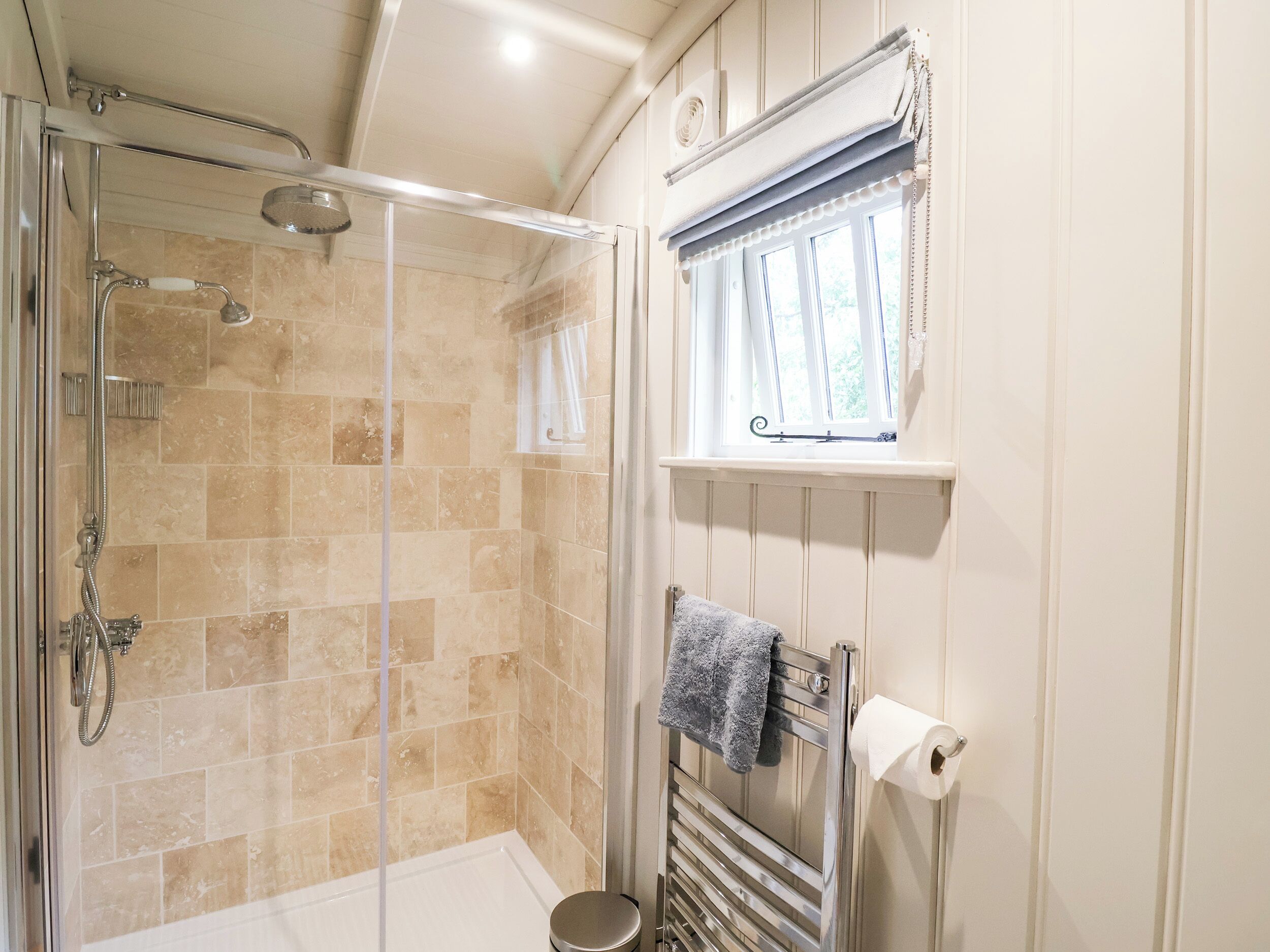 A bathroom with a glass-enclosed shower, beige tiled walls, a towel rack with a towel, and a window with curtains.