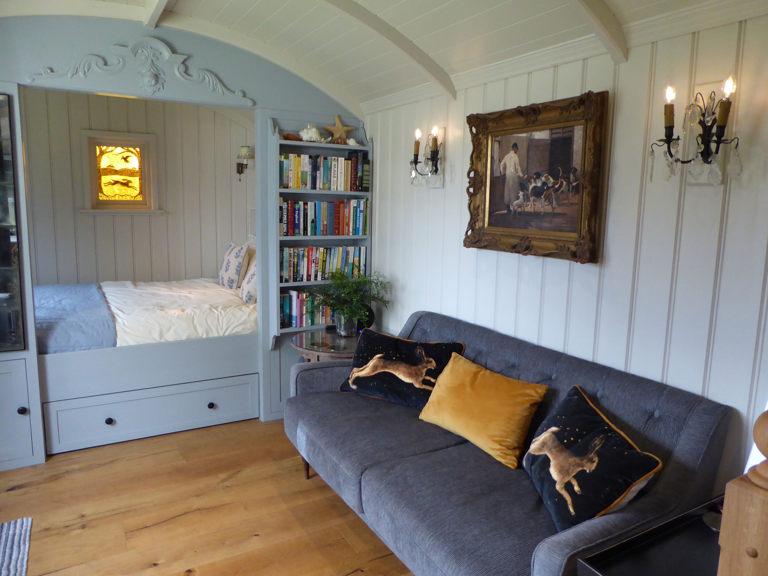 A cozy room with a bed, a grey sofa with decorative pillows, a bookshelf, and a framed picture on the wall.