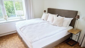 Een opgemaakt bed met witte lakens en een hoofdbord, een nachtkastje met een lamp, en een raam met gordijnen.