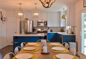 A modern kitchen with white cabinets, a blue island, stainless steel appliances, and a dining area with yellow placemats and white plates. - Salty Sea Shack, Near Park, Beach & Pet-Friendly (Cape Charles)