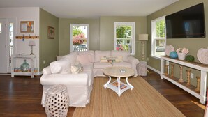 A living room with a beige sofa, a round coffee table, and a TV mounted on the wall. There are windows with a view of the outside, a small side table, and a lamp.