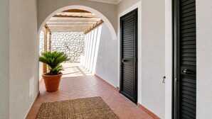 Un pasillo con pared de piedra, una pérgola de madera, una planta en maceta y una puerta negra.