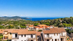 Un pueblo costero con edificios de tejas color terracota, un cielo azul despejado y vistas al mar y montañas distantes.