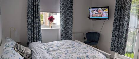 Un dormitorio con cama, una silla, un televisor y una ventana con cortinas.