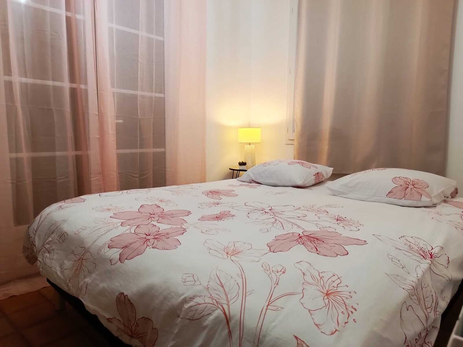 A bedroom with a floral bedspread, a bedside lamp, and sheer curtains.