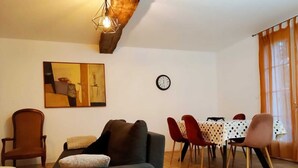 Ein Wohnzimmer mit einer Couch, Sesseln, einem Esstisch mit Stühlen, einer Uhr und einem Gemälde an der Wand.