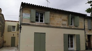 Ein zweistöckiges Gebäude mit beiger Fassade und grünen Fensterläden. Ein Schild ist an dem Gebäude angebracht, und im Erdgeschoss befindet sich ein kleines Fenster.