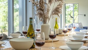 A dining table set with white bowls, wine glasses, and bottles, surrounded by a vase with dried plants and a view of greenery outside. - The Grain Store, Reepham, Norfolk (Reepham)