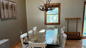 Une salle à manger avec une grande table en bois, des chaises blanches, un lustre et une fenêtre donnant sur les arbres.