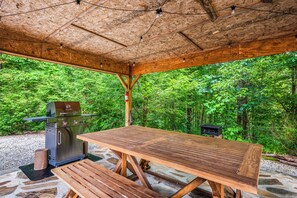 An outdoor wooden pavilion with a grill and picnic table, surrounded by dense forest. - Romantic hot tub cabin | Secluded (Cosby)