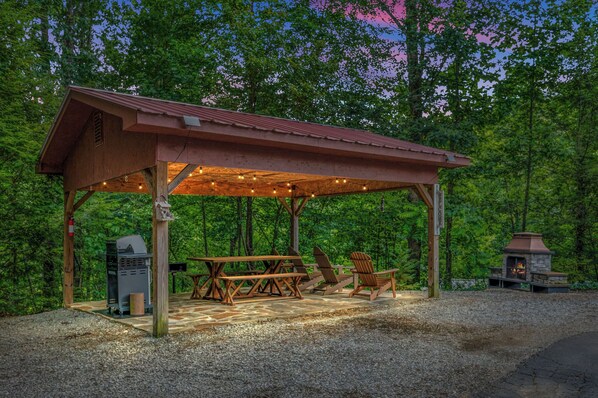A wooden pavilion with a picnic table and chairs, a grill, and a stone fireplace. - Romantic hot tub cabin | Secluded (Cosby)