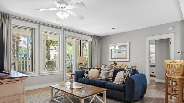 A living room with a blue sofa, wooden coffee table, and large windows. - Poolside 2BR Condo at Baytowne Wharf! GOLF CART to Beach, Pool, Hot Tub, Tram (Miramar Beach)
