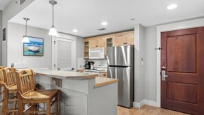 A kitchen with a central island, wooden chairs, a stainless steel refrigerator, and a wooden door. - Poolside 2BR Condo at Baytowne Wharf! GOLF CART to Beach, Pool, Hot Tub, Tram (Miramar Beach)