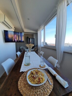 A dining area with a long table, white chairs, a TV mounted on the wall, and a view of the city through large windows. - Penthouse F8 | Rooftop Terrace, Two Towers View (Bologna)