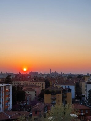 A cityscape at sunset with buildings in the foreground and a clear view of the sun setting behind the skyline. - Penthouse F8 | Rooftop Terrace, Two Towers View (Bologna)