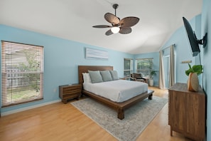 A bedroom with a bed, wooden headboard, bedside tables, a ceiling fan, a TV mounted on the wall, and a window with blinds.