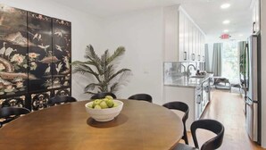 A modern kitchen with a dining area, featuring a large wooden table with a bowl of apples, a potted plant, and a decorative screen with a nature scene.